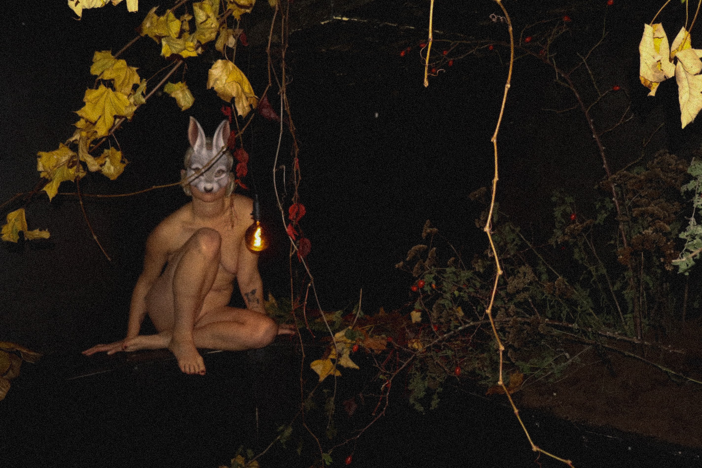 Rabbit-masked performer sitting among branches and leaves in Prey for Me